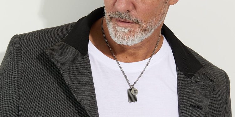 Mann trägt eine silberne Halskette mit zwei markanten Anhängern, kombiniert mit weißem T-Shirt und dunklem Blazer