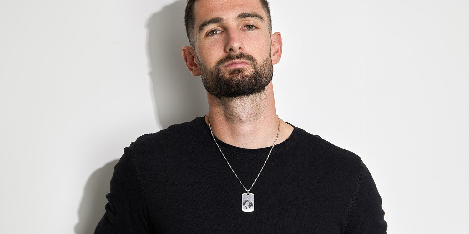 Mann mit Bart trägt eine silberne Halskette mit rechteckigem Anhänger, gekleidet in schwarzem T-Shirt vor weißem Hintergrund