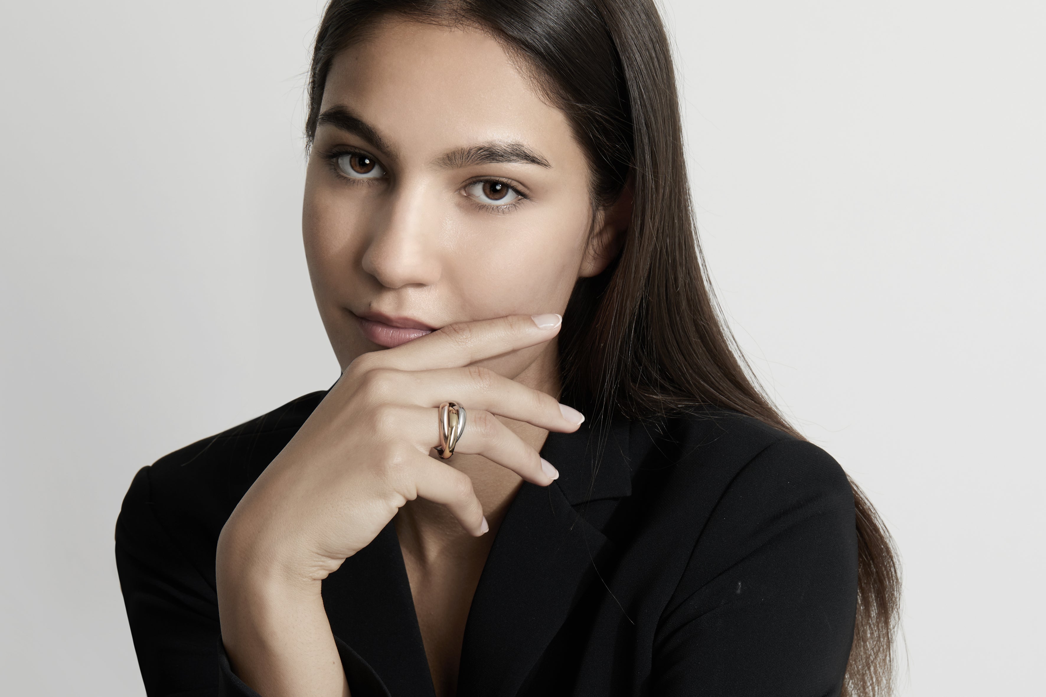 Frau mit dunklen Haaren in schwarzem Blazer hält nachdenklich die Hand am Kinn und präsentiert einen eleganten goldenen Ring vor hellem Hintergrund