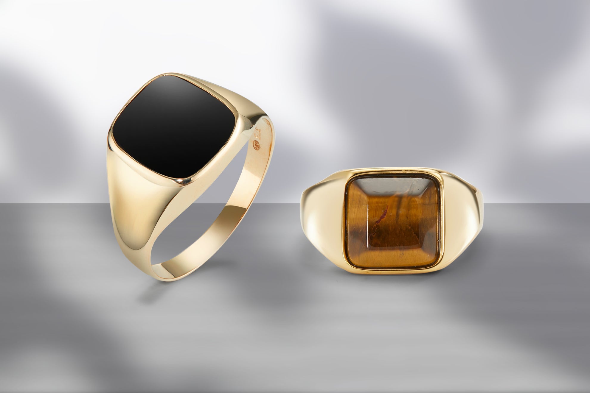 Zwei goldene Herren-Siegelringe mit schwarzem Onyx und braunem Tigerauge in klassischem Design vor grauem Hintergrund