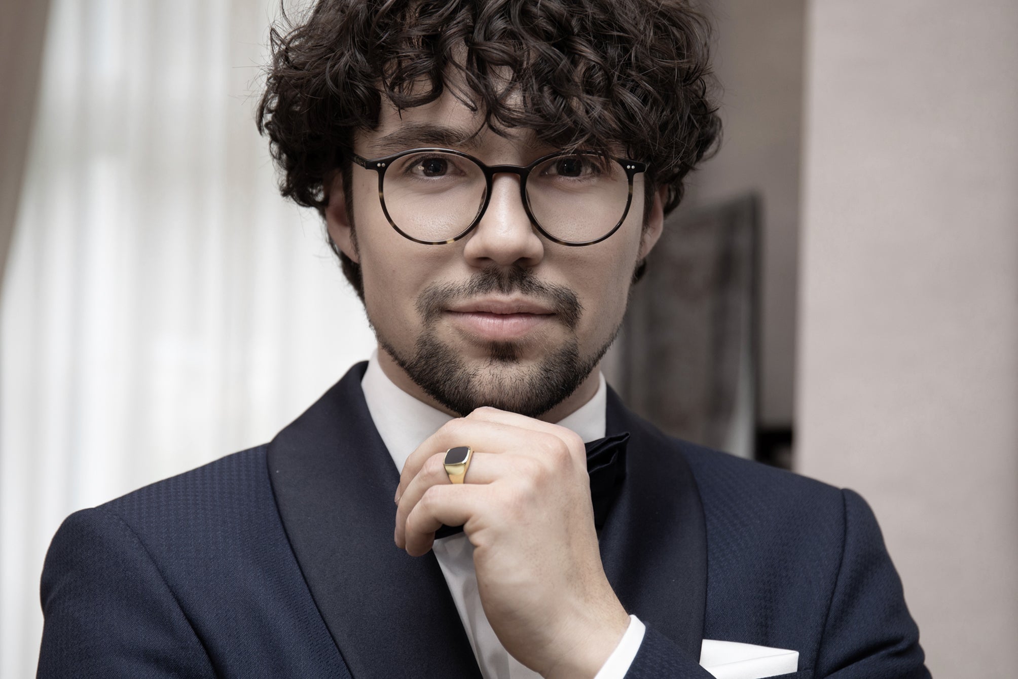 Eleganter Mann mit lockigen Haaren und Brille trägt einen goldenen Siegelring zu dunklem Anzug und weißem Hemd in stilvollem Ambiente
